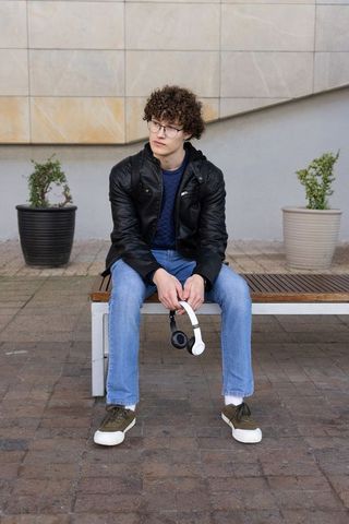 Young man sitting on bench holding headphones looking thoughtful in urban plaza