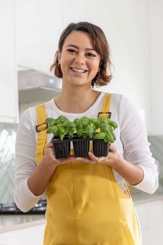 Smiling african american woman holding basil seedling tray in kitchen wearing mustard overalls