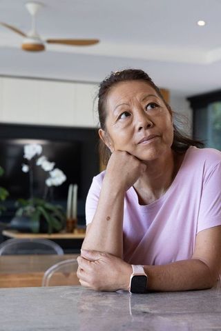 Asian senior woman contemplating at kitchen counter wearing pink tee and smartwatch