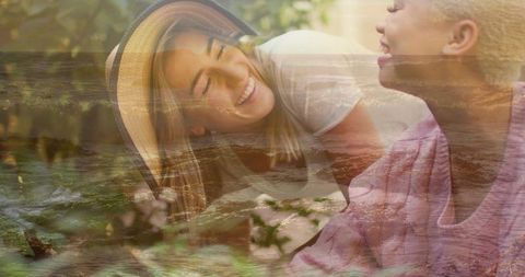 Smiling women sharing moment, leaning close on sunlit patio, double-exposure ocean overlay
