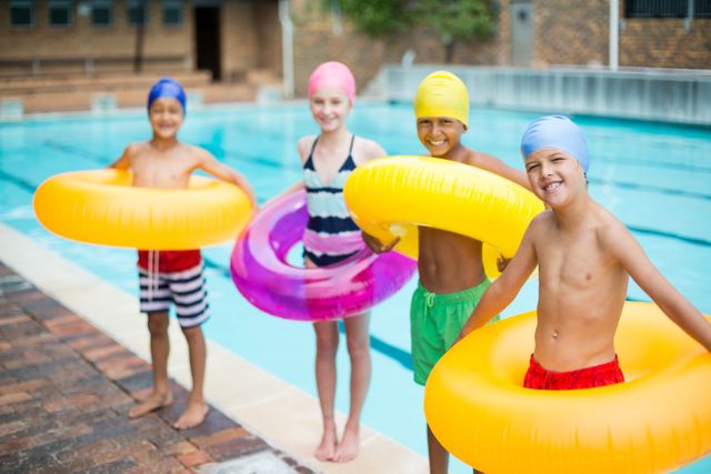 Children standing at poolside holding colorful inflatable rings, wearing swim caps and swimwear. Perfect for use in summer vacation promotions, swimming pool advertisements, children's activity brochures, and family fun campaigns.