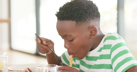 Young boy eating noodles with fork at bright sunlit kitchen table, casual mealtime