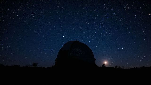 Observatory dome silhouetting against vast starfield with rising moon and distant trees