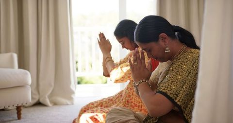 Indian mother and daughter engage in meditation, wearing traditional sarees in a peaceful domestic setting. Light filters gently through curtains, creating a sense of tranquility and inward reflection. Useful for themes about Indian culture, family traditions, spiritual practices, mindfulness, and inner peace. Ideal for promoting cultural awareness, meditation and wellness platforms, or illustrating articles about family bonding and cultural customs.