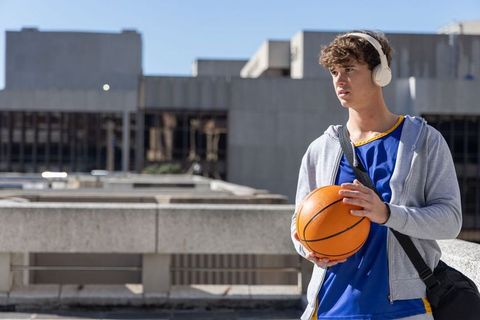 Teen basketball player holding ball on urban rooftop wearing headphones and jersey