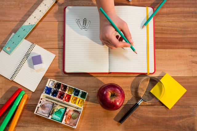 Hand writing in notebook on wooden table with various school supplies. Ideal for educational content, back to school promotions, and creative learning materials.