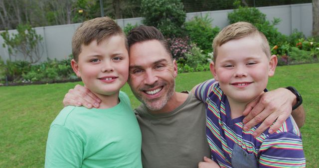 Man smiling and embracing two young boys in a backyard, displaying family bonding and happiness. Ideal for advertising family-oriented products, parenting advice articles, or family event promotions.