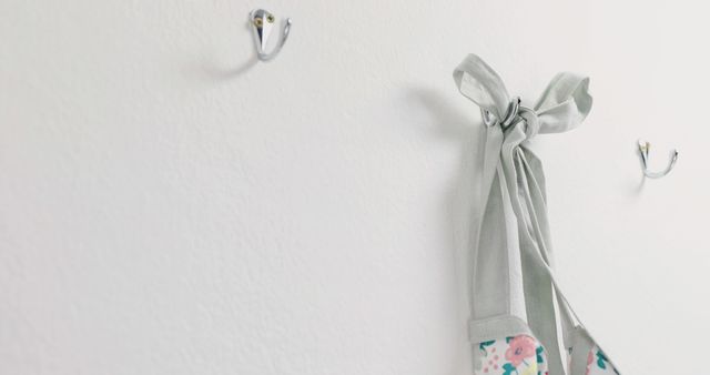 Floral apron hanging on metal wall hooks. Clean, minimalist white wall with one apron positioned to the right side. Perfect for content about home organization, interior design, kitchen decor, or minimalist living.