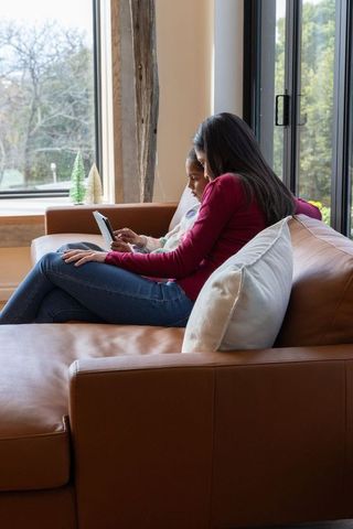 African american mother and daughter sharing tablet on brown leather sofa near window