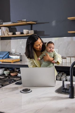 Mother holding infant while working on laptop at modern kitchen island, multitasking caregiving mome
