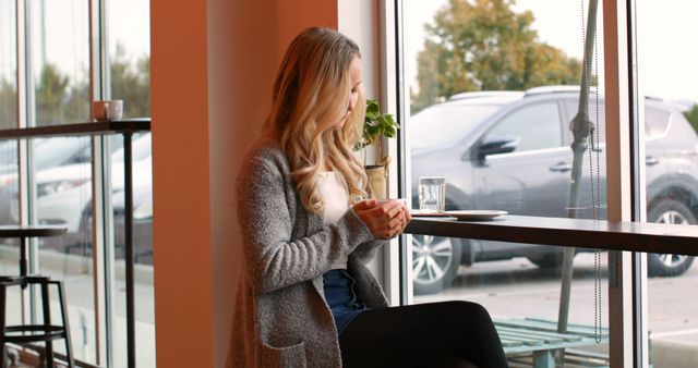 Young woman sitting alone by window in a cozy cafe, enjoying her coffee. Perfect for content related to cafes, leisure, relaxation, morning routines, and lifestyle blogs or articles. Ideal for marketing campaigns promoting cafes, coffee shops, or relaxation products.