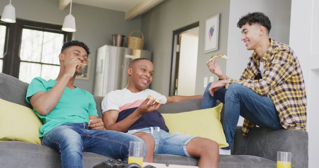 Three young men, casually dressed, are sitting on a grey sofa in a modern living room. They are eating pizza and laughing together. Bright pillows add a pop of color to the scene, along with joy and camaraderie suitable for promoting lifestyles, youth culture, friendship, and leisure activities.