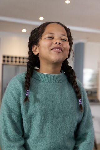 Young girl laughing, standing in cozy kitchen wearing teal sweater with purple hair ties