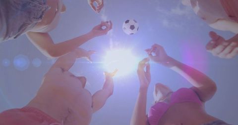 Friends reaching for soccer ball under bright sun at beach, summer play and joyful vibes