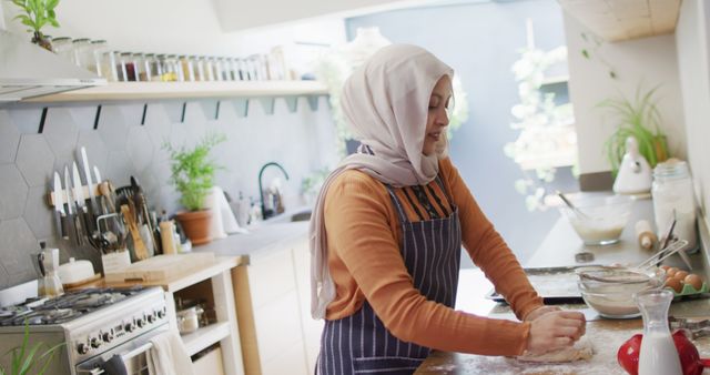 Image of happy biracial woman in hijab baking in kitchen at home, kneading dough. Happiness, health, inclusivity and domestic life.