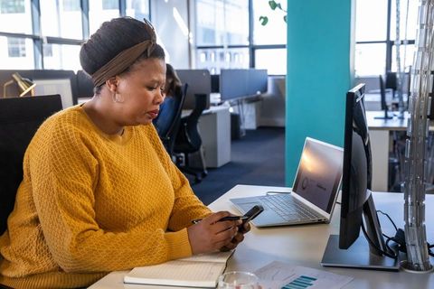 African american professional checking smartphone while working at desk with laptop and monitor