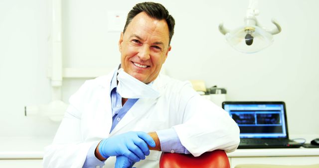 Smiling dentist standing in a modern dental office, exuding confidence and friendliness. The dental professional is wearing a white lab coat, gloves, and has a mask lowered to reveal a warm smile. Background includes dental equipment and a computer screen with dental X-rays. Ideal for promoting dental clinics, healthcare services, and medical professionals.