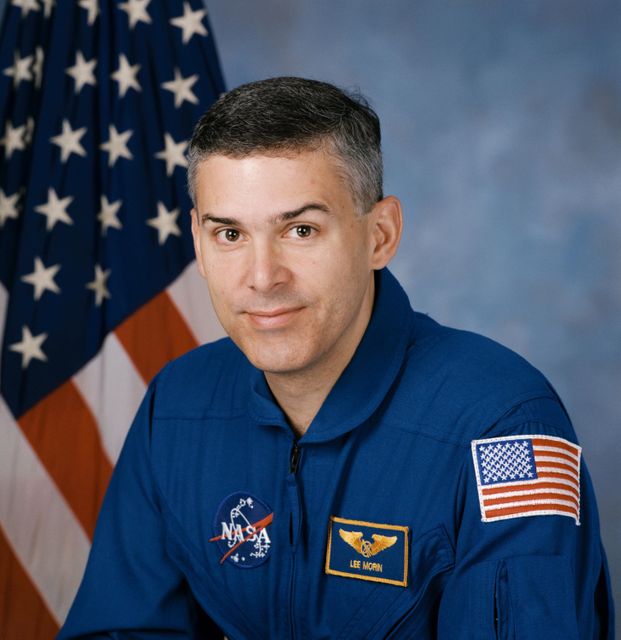 In this image, an astronaut dressed in a blue NASA uniform is posing in front of the American flag. The patch on his arm signifies his role as a mission specialist. Such a photograph can be used in articles, educational materials, or presentations about space exploration, NASA missions, and the achievements of astronauts. This official portrait is ideal for highlighting patriotism, dedication, and careers in aerospace.