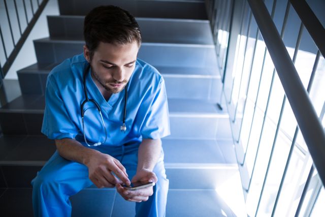 Male nurse in blue scrubs and stethoscope sitting on staircase in hospital, using mobile phone. Ideal for use in healthcare, medical, and technology-related content. Perfect for illustrating themes of healthcare professionals, work breaks, communication in medical settings, and the use of technology in healthcare.