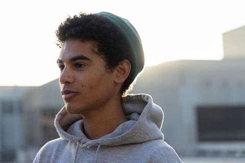 Backlit urban teen standing on rooftop wearing gray hoodie and green beanie during golden hour