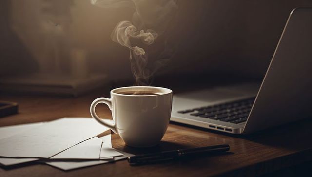 Steaming white ceramic mug resting on wooden desk with laptop and paperwork, cozy workspace