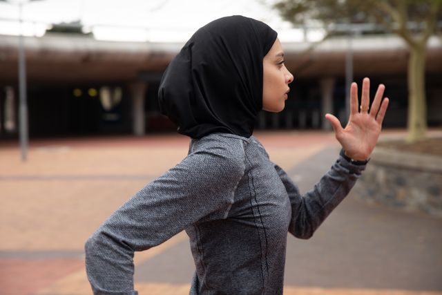 Fit biracial woman wearing hijab and sportswear exercising outdoors in the city, running in urban park. Urban lifestyle exercise.