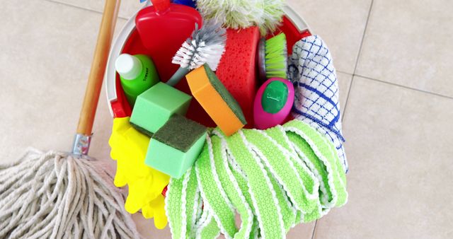 Brightly colored cleaning supplies arranged in bucket, including various sponges, brushes, cloths, and bottles on light tile floor. Ideal for illustrating household chores, cleaning routines, or advertising cleaning products and services. Useful for blog posts, articles, or marketing materials focused on cleanliness and hygiene.