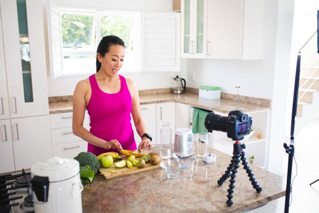 Asian female vlogger slicing fruits to make healthy juice in a modern kitchen. Ideal for content related to healthy lifestyles, cooking vlogs, influencer marketing, and technology in everyday life. Suitable for promoting kitchen appliances, healthy eating habits, and fitness routines.