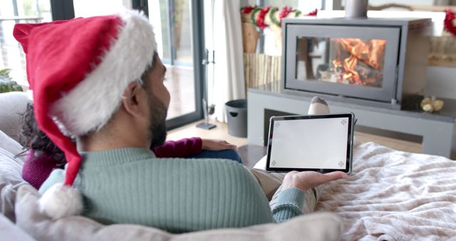 African American man wearing Santa hat using tablet by fireplace during Christmas. Suitable for themes of holiday celebrations, cozy home settings, digital technology use during festive seasons, and relaxed indoor environments.