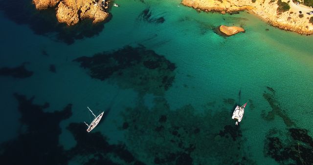 Aerial perspective showing two sailboats anchored in clear turquoise waters close to a rocky coastline. Ideal for use in travel blogs, tourism promotions, marine-related publications, and advertisements for sailing or vacation destinations.