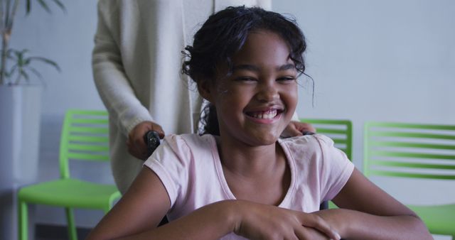 Happy African American girl in wheelchair, smiling at camera, assisted by a caregiver from behind. Ideal for topics on health care, support, and inclusion. Suitable for use in medical and educational contexts, promoting awareness and positivity.