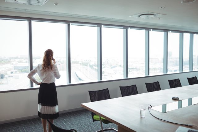 Businesswoman standing in a modern boardroom, talking on her mobile phone while looking out the window at a city view. Ideal for use in business, corporate, and professional contexts, such as websites, presentations, and marketing materials highlighting professional environments and executive communication.