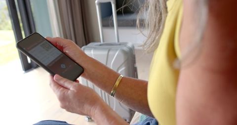 Mature woman uses smartphone for travel booking with suitcase