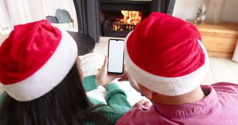 Couple wearing santa hats sharing smartphone by fireplace, cozy holiday evening