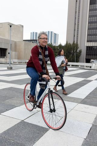 Mature man riding fixed-gear bicycle across urban plaza while woman walks with travel mug
