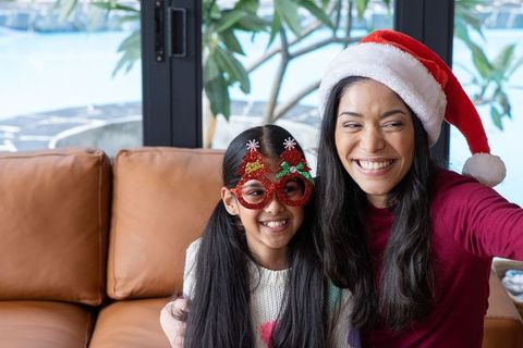 Mother and daughter taking christmas selfie wearing santa hat and tree glasses on couch