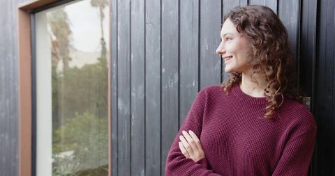 Young woman leaning on dark wood wall wearing maroon sweater smiling looking right