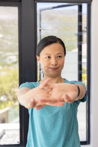 Mature asian woman stretching arms forward in teal shirt wearing smartwatch at home