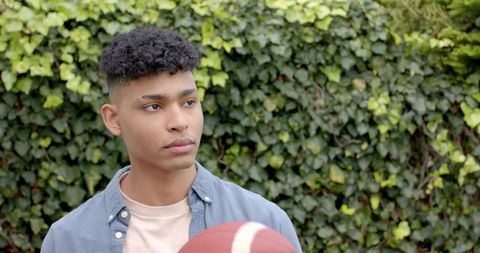Young man standing in garden holding football looking aside against ivy-covered wall