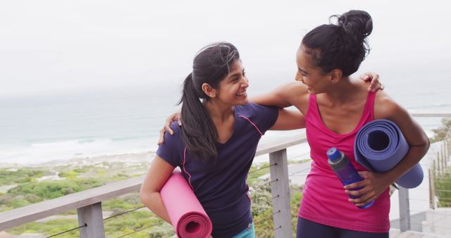 Two women are bonding after a yoga session by the coast, smiling and holding yoga mats. This stock photo is perfect for promoting fitness, wellness, outdoor activities, and friendship.