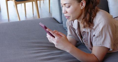 Young woman reclining on gray sofa scrolling smartphone, casual living room lifestyle daylight