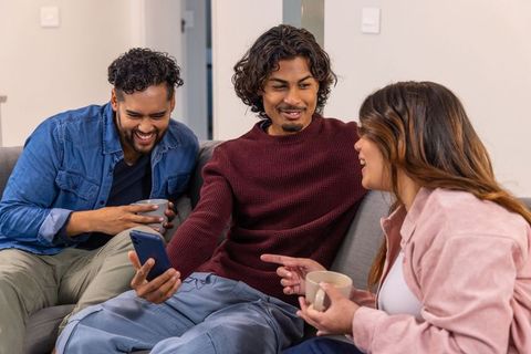 Asian friends laughing together on sofa, sharing smartphone and coffee at casual hangout