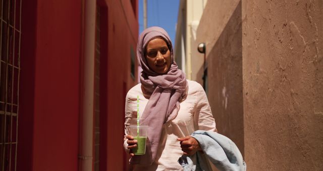 Young woman in pink hijab enjoying a green smoothie while walking in an alleyway on a sunny day. This image is perfect for content related to urban lifestyle, health and wellness, diversity, and fashion.