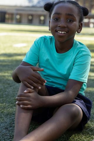 African american girl sitting on grass smiling wearing teal t-shirt navy shorts park