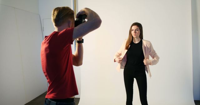 Photographer taking photo of young model in a studio with white background. Model wearing casual clothes, adjusting jacket, and ready for the shot. Can be used for themes related to fashion photography, modeling, studio setup, and professional photography sessions.