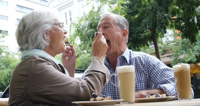 Elderly couple sitting at a cafe table outside, feeding each other dessert and enjoying lattes. The green park environment and bright day enhance the relaxed and happy moment shared between the senior citizens. Great for portraying themes of companionship, retirement, happy living, and the pleasure of simple moments in promotional or editorial use.