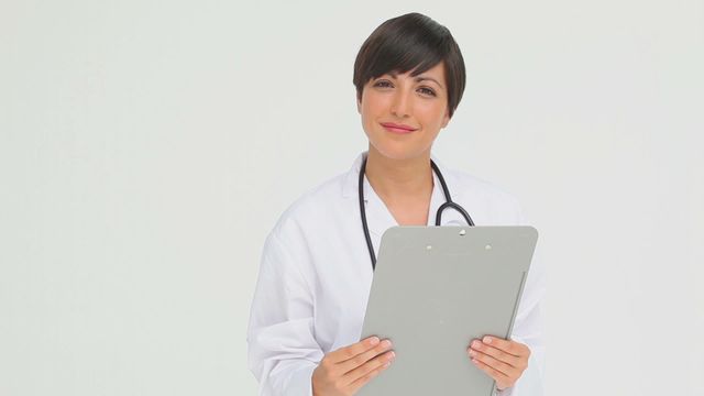 Medical professional standing against a grey background, holding a clipboard and wearing a white coat. Intent on emphasizing sincerity and approachability. Ideal for healthcare, medical ads, and brochures relating to medical services or careers.