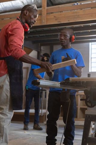 African american instructor teaching apprentice woodworking using table saw in workshop