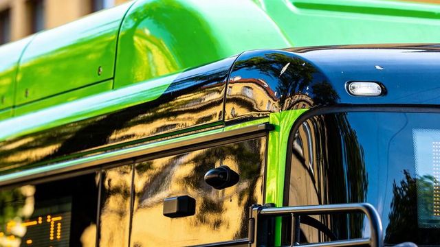 Glossy green-black city bus front corner reflecting urban facades and trees in sunlight