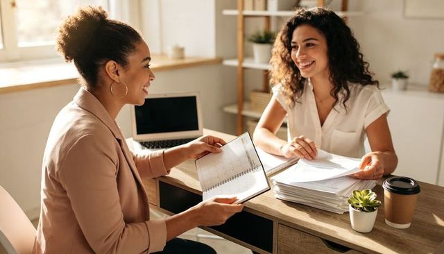 Two women collaborating over planner and documents, smiling teamwork with laptop and coffee